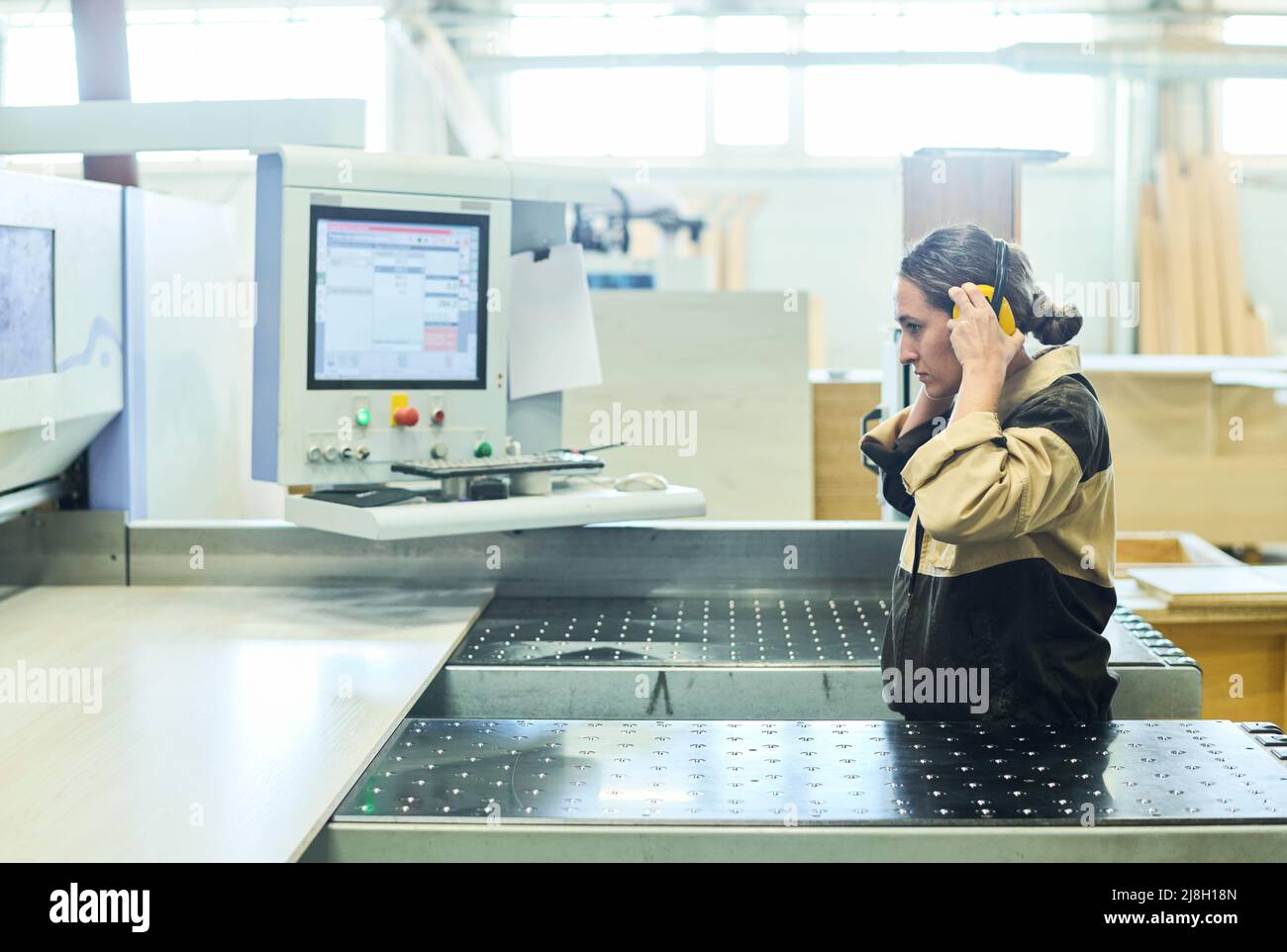 Female worker wearing protective headphones while working at manufacturing equipment with wooden board Stock Photo