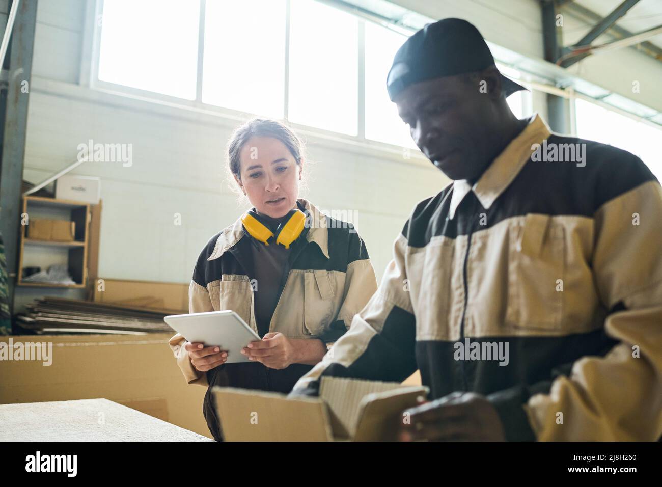 Young woman in uniform using digital tablet while her colleague packing box at table Stock Photo