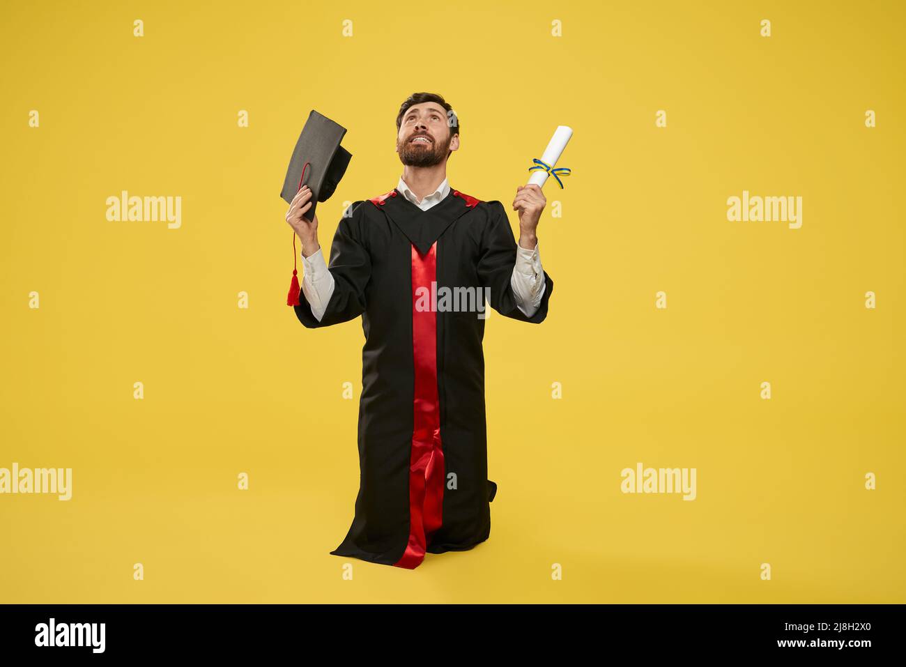 Frint view of brunette student wearing graduate gown standing on knees. Young male holding diploma and mortarboard, looking up, happy, glad, grateful. Stock Photo