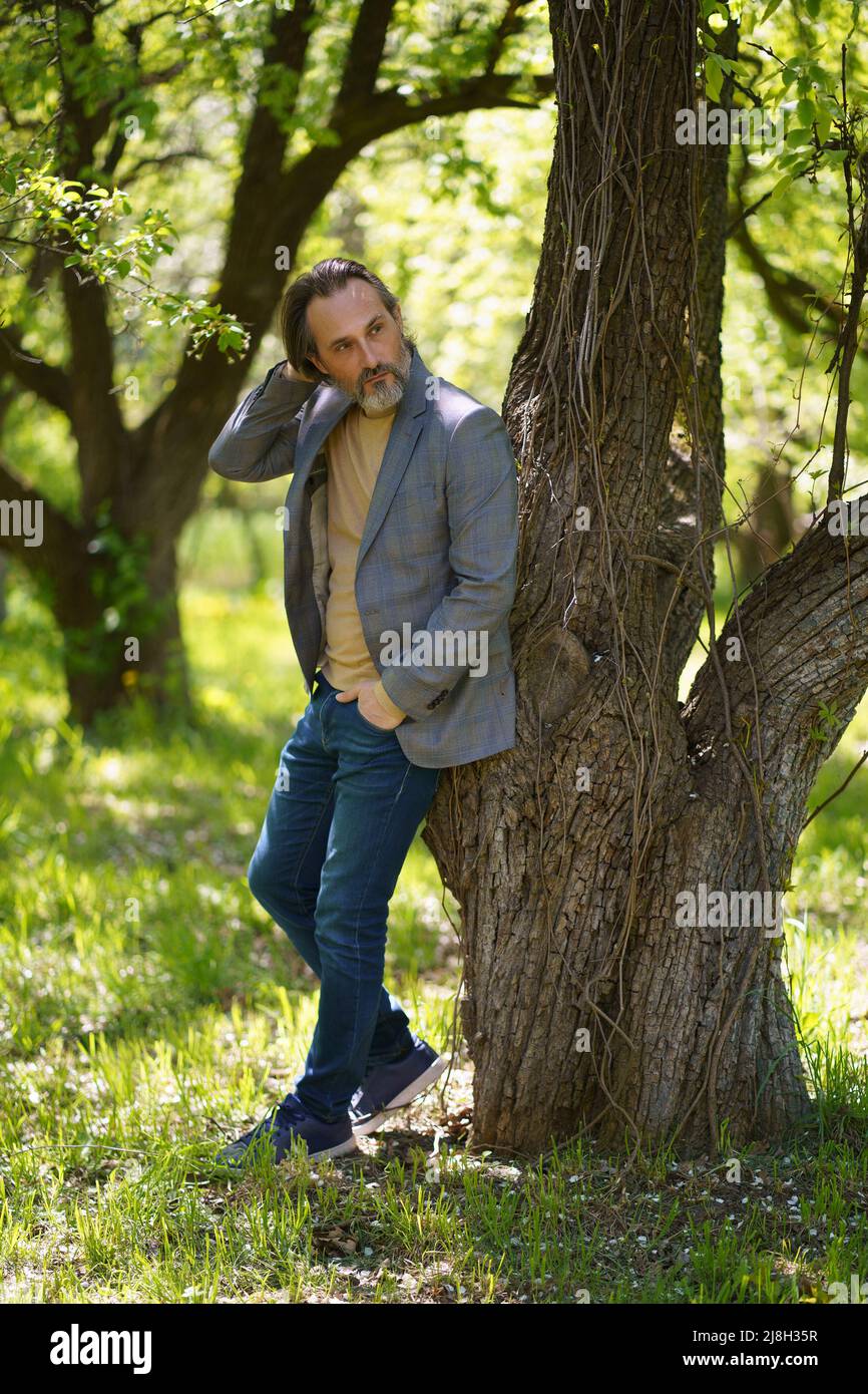 A nice looking mature birded man in casual standing under tree on the grass looking behind the tree with hands in pockets. Freelancer spend time working outdoors.  Stock Photo