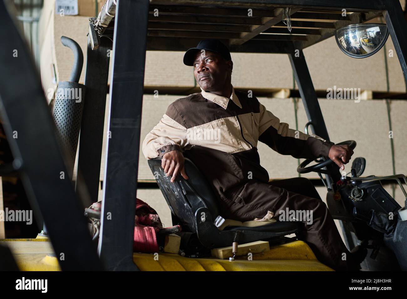 African male worker in uniform driving forklift and transporting freight in warehouse Stock Photo