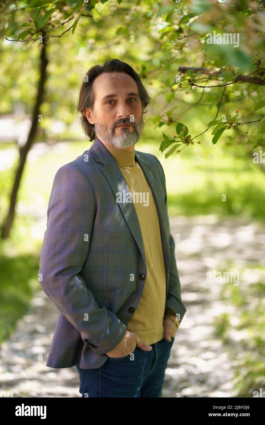 Mature handsome man with grey beard looking thoughtful at the camera wearing casual jacket and jeans holding hands in pockets.  Stock Photo