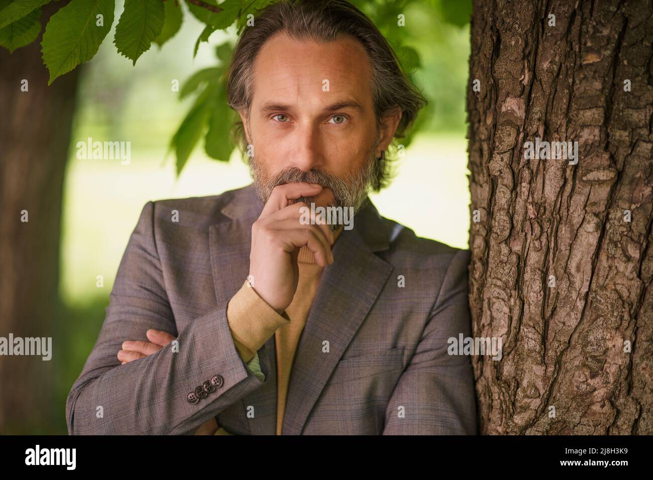 Charming mature grey birded businessman in casual standing under tree on the grass looking at camera with arms folded touching his face. Freelancer spend time working outdoors.  Stock Photo