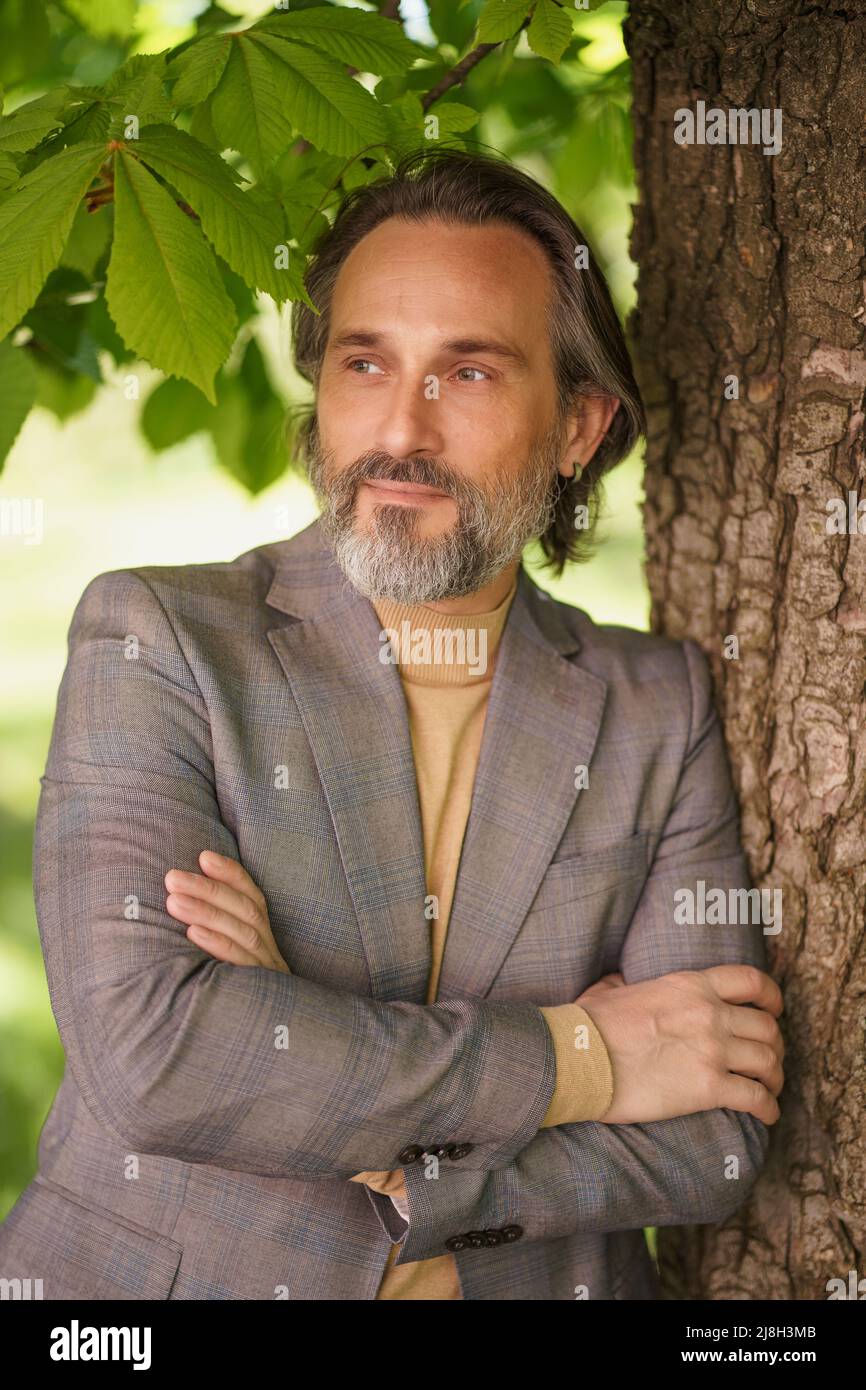 Handsome mature grey birded man in casual standing under tree on the grass looking away with arms folded. Freelancer spend time working outdoors.  Stock Photo