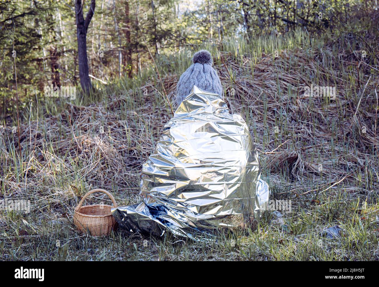 Woman person is lost and sit in wild forest in cold day, using first aid emergency blanket to prevent hypothermia and body heat loss. Stock Photo