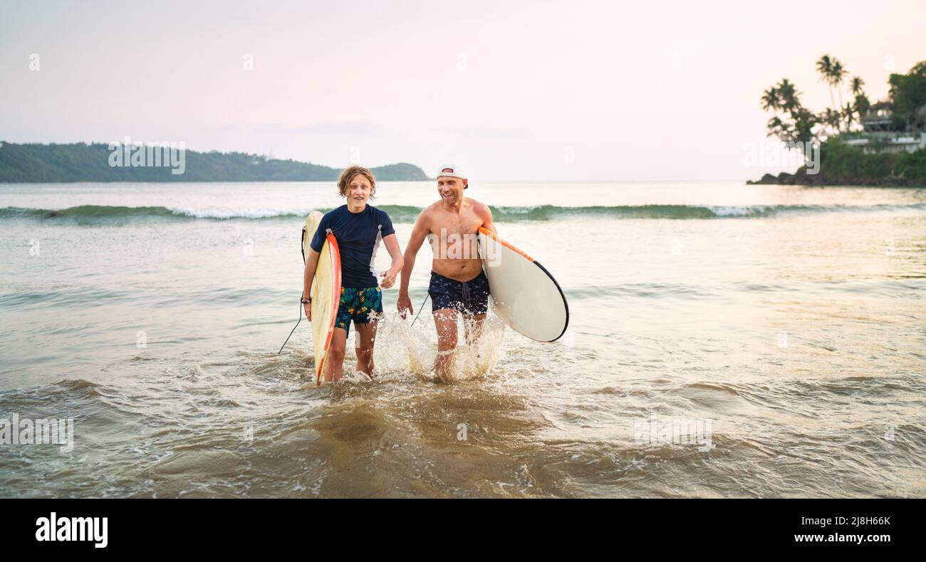 Portrait of a teen boy with his father with surfboards after surfing. They are smiling and walking out of the water. Family active vacation or active Stock Photo