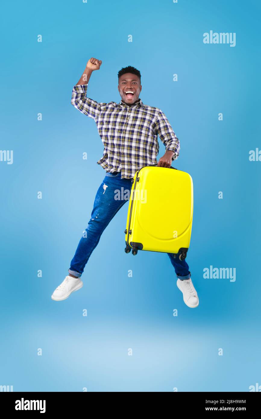 Excited young African American guy with yellow suitcase jumping and gesturing YES on blue studio