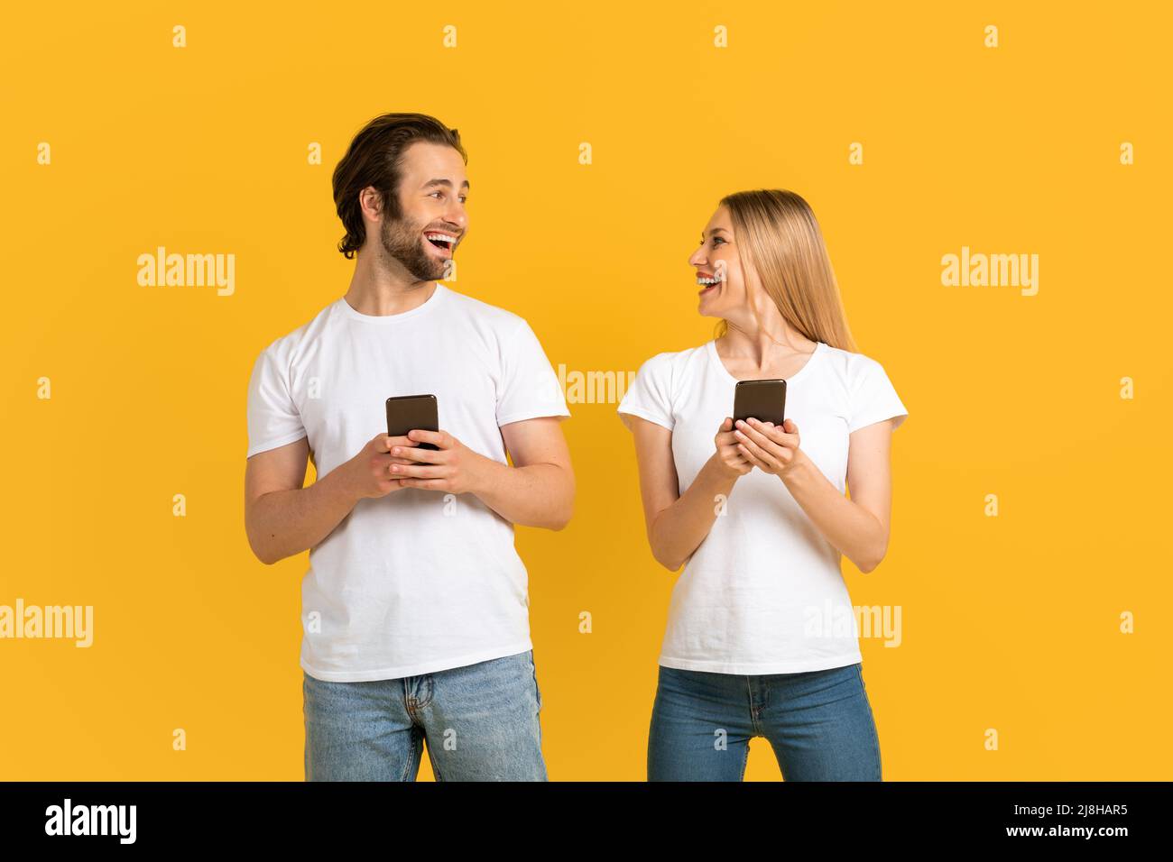 Cheerful millennial caucasian female and male in white t-shirts look at each other, hold phones Stock Photo