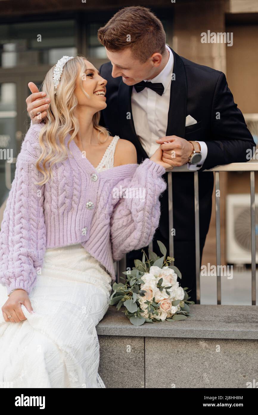 Beautiful young wedding couple in black suit and white dress walk around city, laugh and hug on street background. Portrait of happy bride with Stock Photo