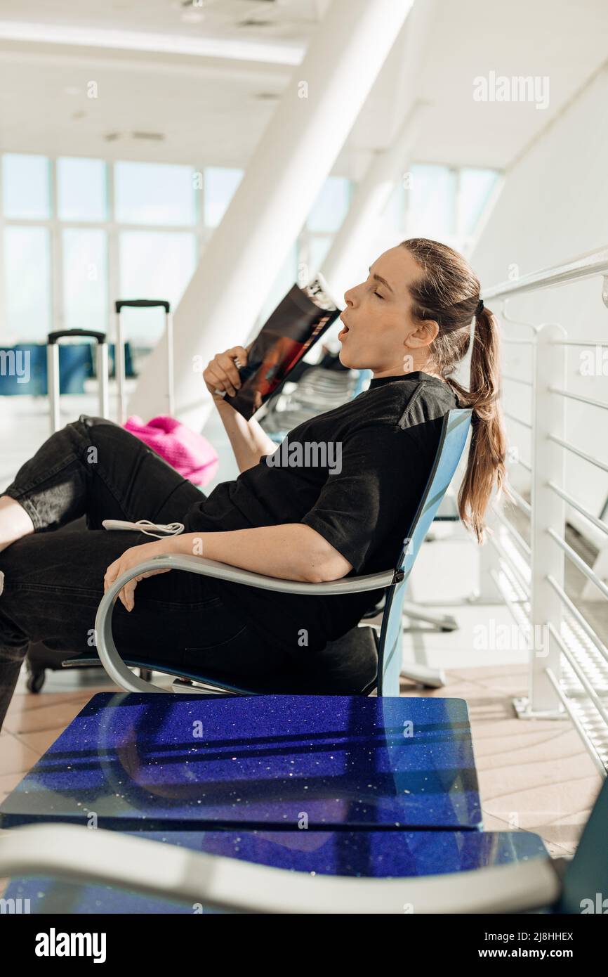 Young woman sitting on chair in airport near suitcases with clothing, suffering from heat, yawning and waving magazine while waiting for airplane Stock Photo