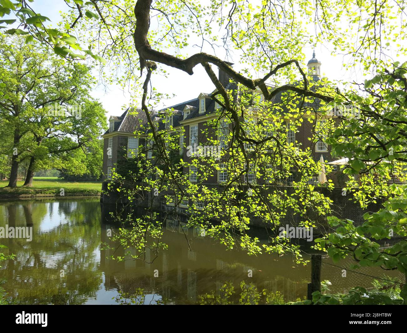 View of the moated manor house of De Wiersse, from across the stream with dappled sunlight and spring foliage; Vorden, the Netherlands, May 2022 Stock Photo