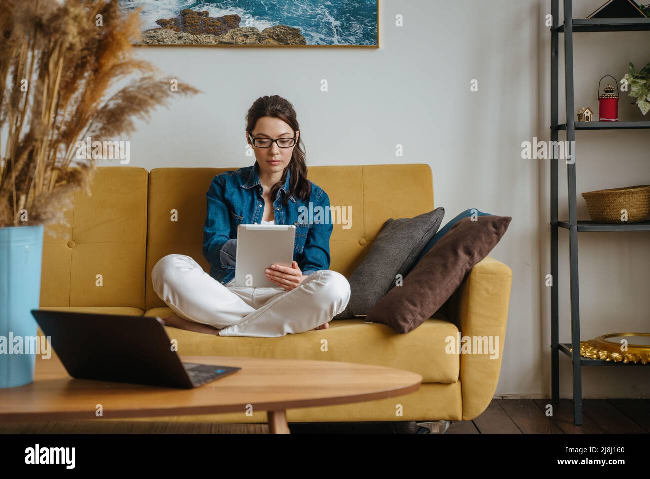 Happy woman reading a book in an ebook reader sitting on a couch at home Stock Photo