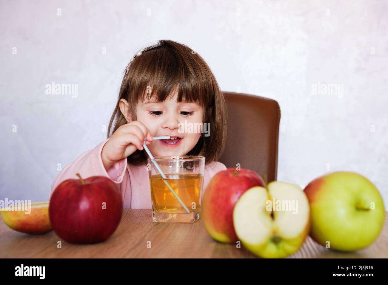Cute little girl drinks apple juice using drinking straw. Baby girl with juice and fruit apples. Stock Photo