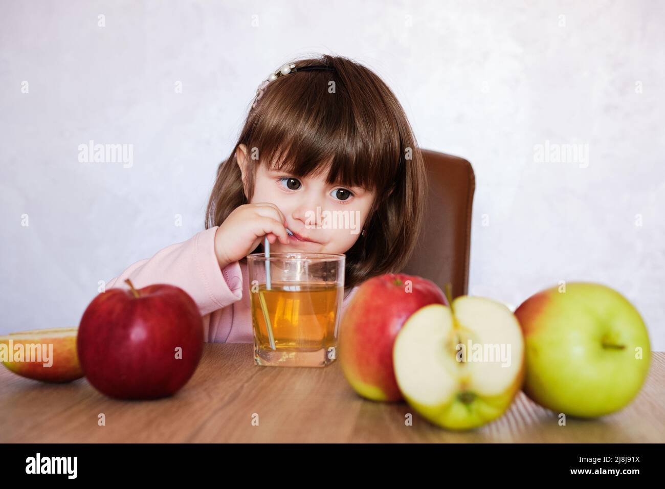 Cute little girl drinks apple juice using drinking straw. Baby girl with juice and fruit apples. Stock Photo