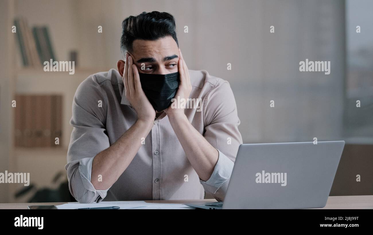Bored tired hispanic guy worker in medical mask during pandemic coronavirus young business man tired ill worker feel boredom in home office hold head Stock Photo