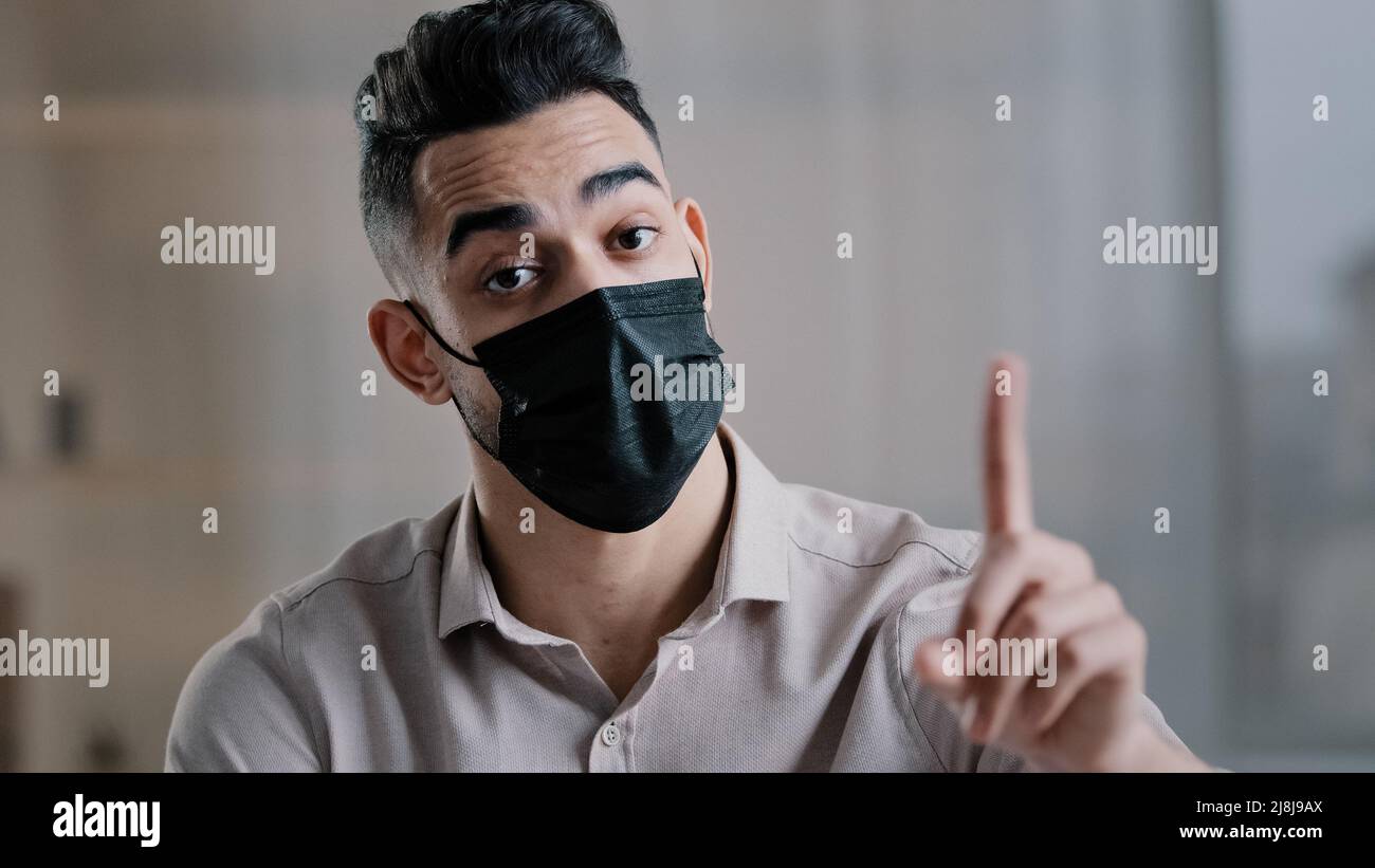 Arabian businessman employee wear medical mask sit at office desk shakes wave index finger warn illnesses infection showing no gesture disagreement Stock Photo
