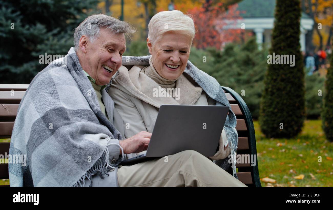 Older happy caucasian husband wife mature elderly couple grandparents family look at screen laptop work remotely browser good news watch funny video Stock Photo