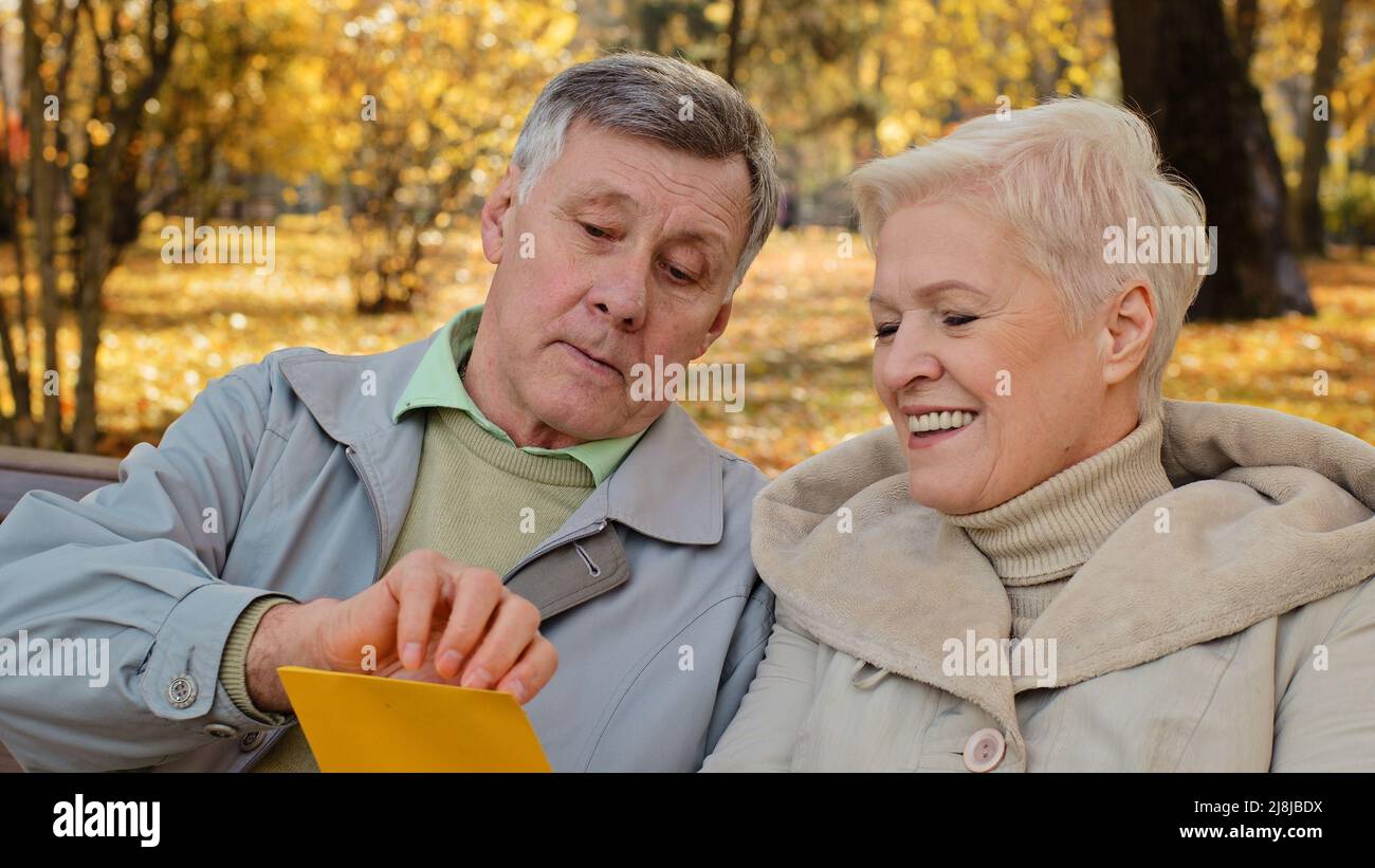 Smiling elderly couple aged partners grandparents senior wife husband open incoming postal envelope reads good news mail letter bank loan paper Stock Photo