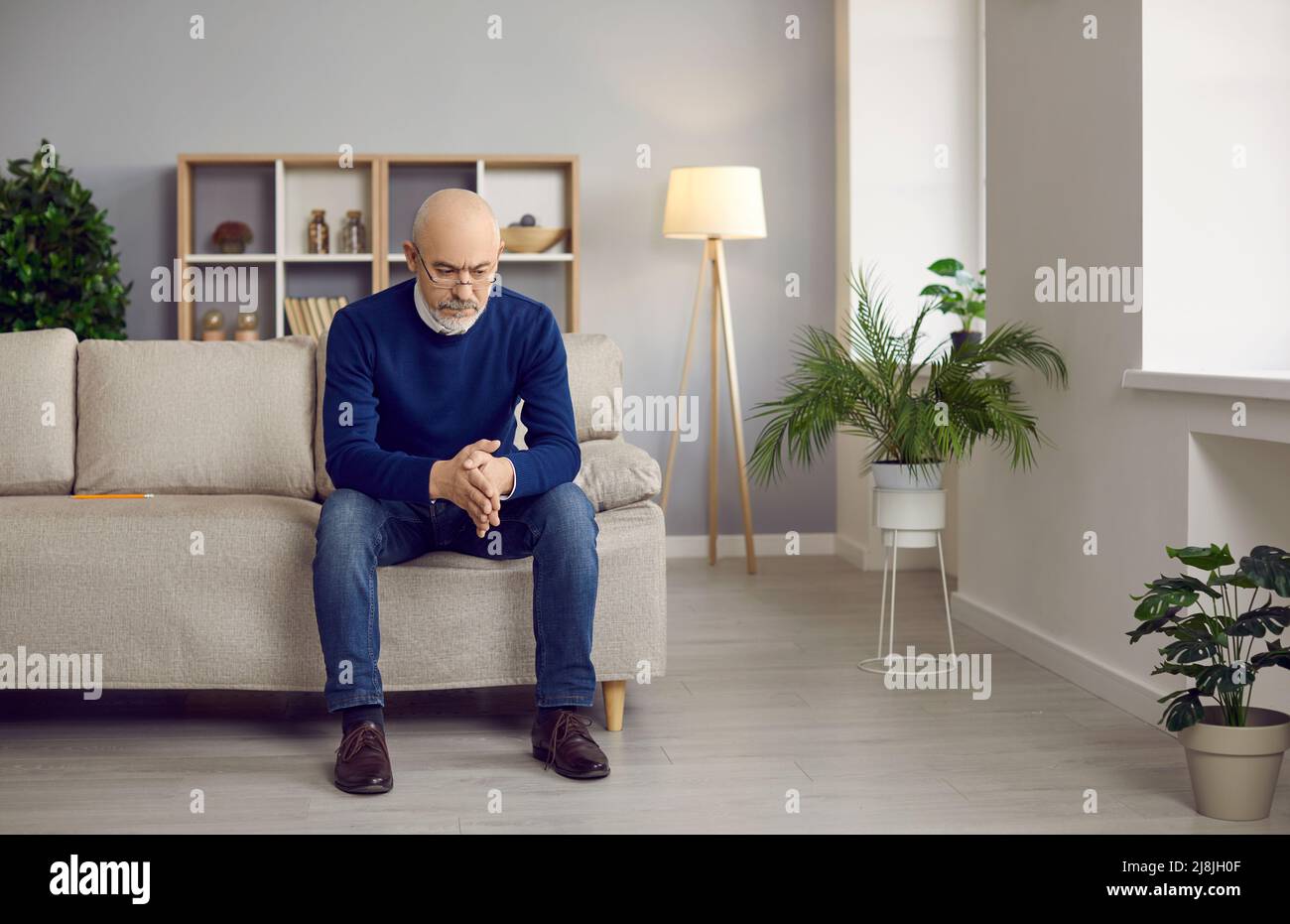 Older man with serious expression sits on sofa at home and thinks about his life and business. Stock Photo