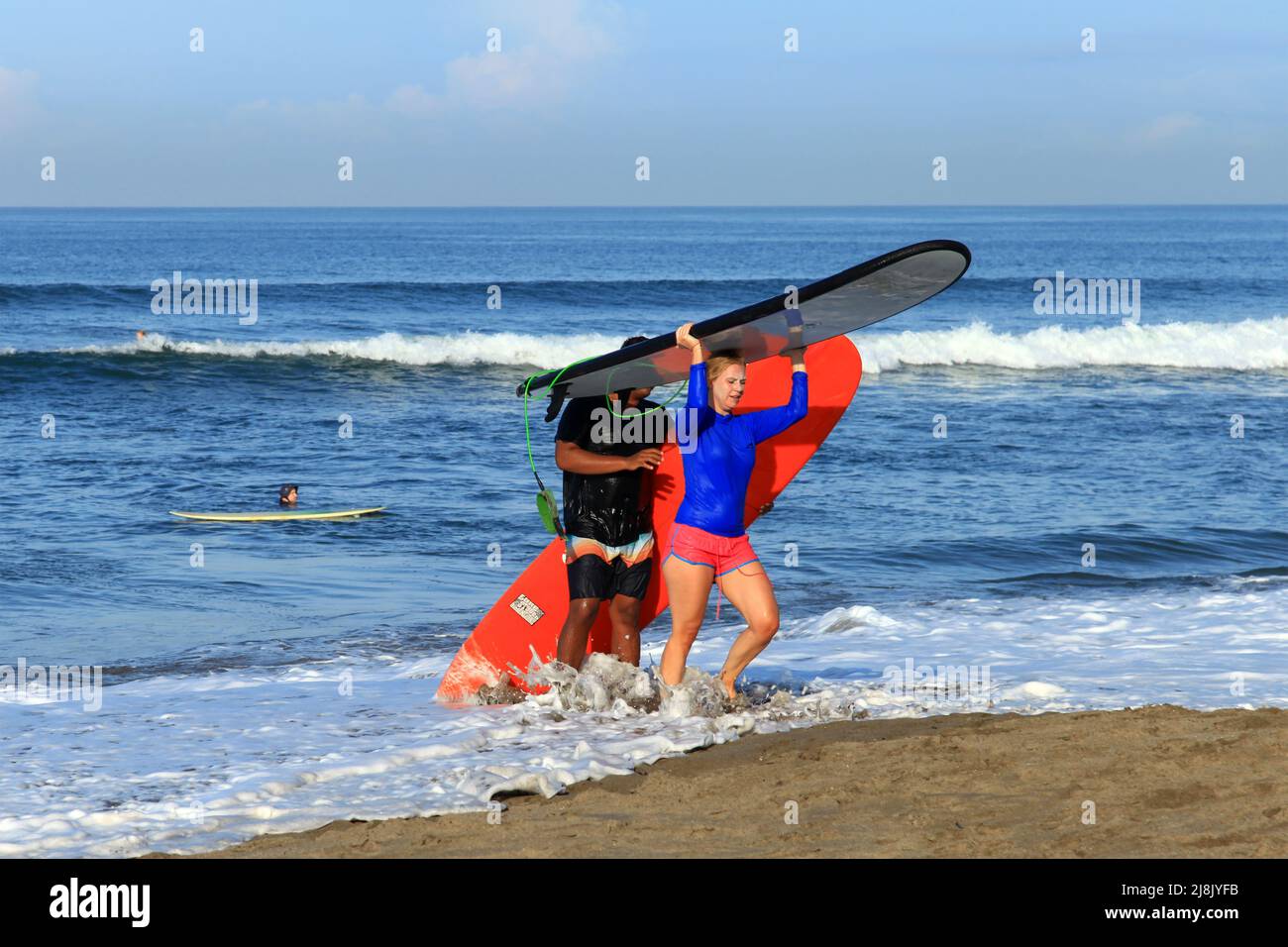 A blonde Caucasian woman with her surf instructor carrying longboards