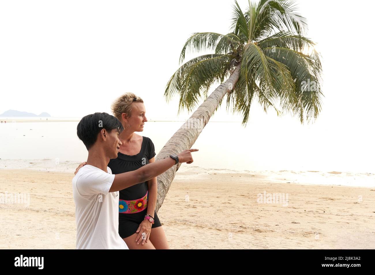 Young asian man pointing ahead next to a woman sitting on a palm tree Stock Photo