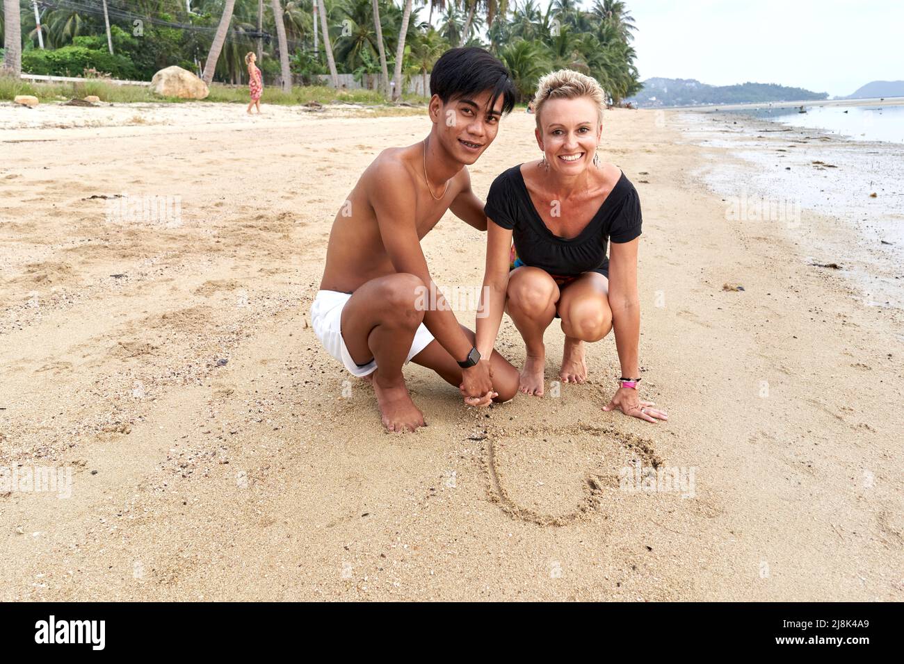 Couple crouching on the beach while drawing a heart shape on the sand Stock Photo