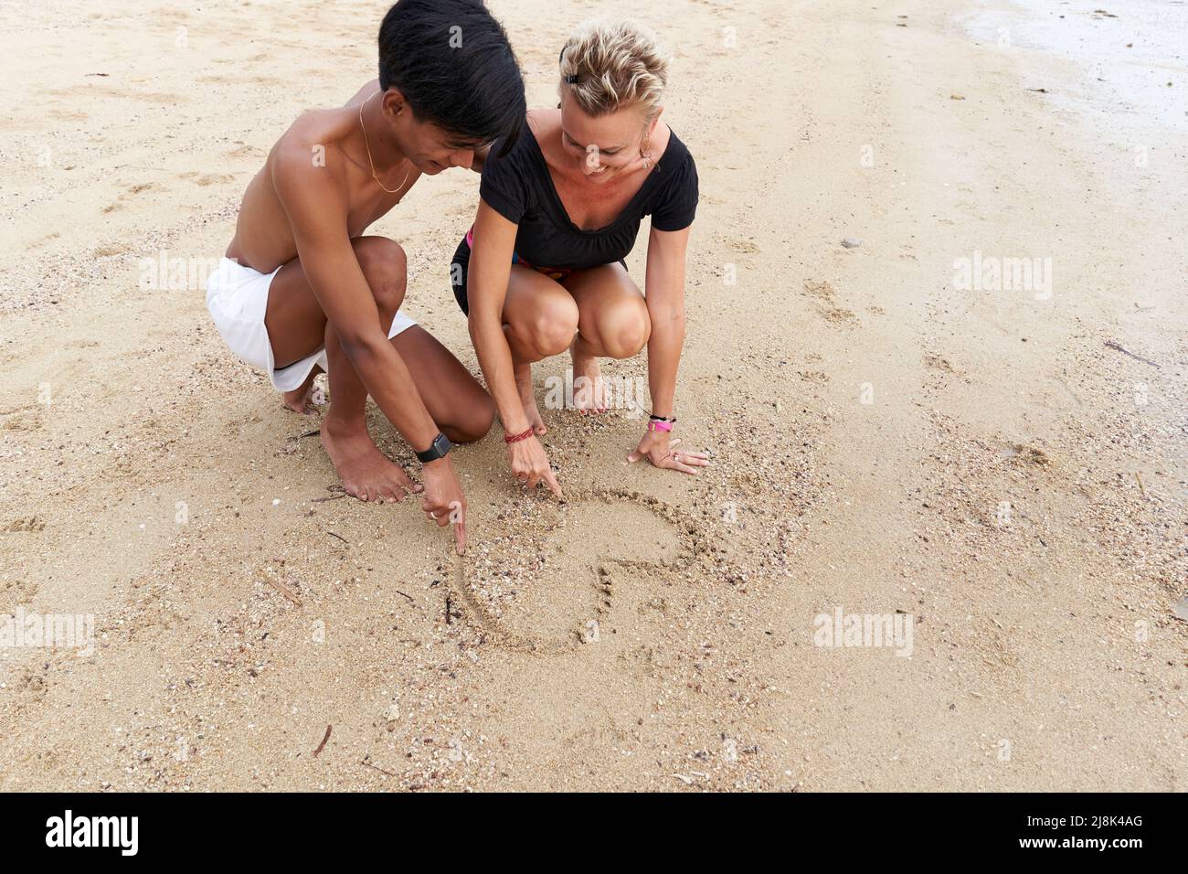 Multiracial couple drawing a heart on the sand of a tropical beach Stock Photo