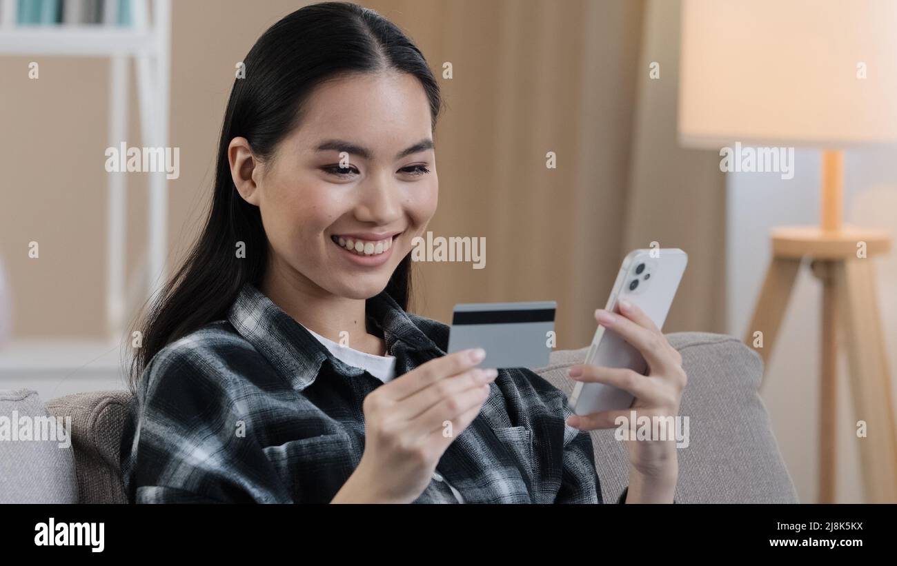 Smiling asian ethnic woman customer user holding bank credit card with virtual money typing smartphone at home. Happy multiethnic female shopper using Stock Photo