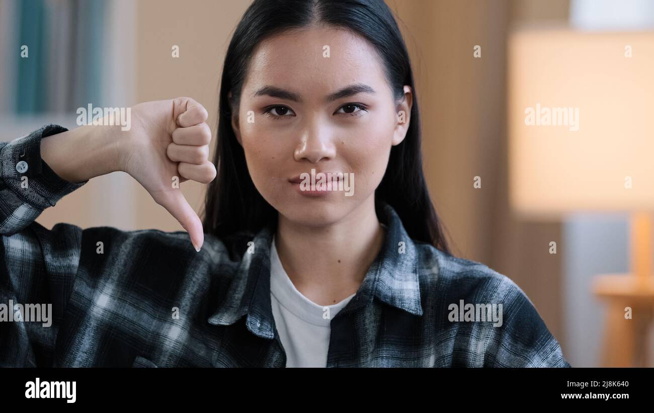 Close up asian millennial woman korean girl wear casual plaid shirt looking at camera showing dislike thumb down disagreement disgust rejection sign Stock Photo