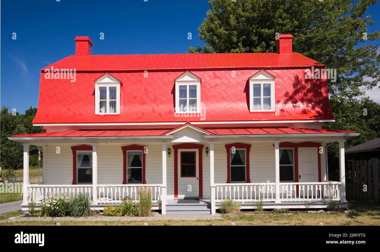 Old circa 1825 Canadiana cottage style home with white wood plank cladding and red sheet metal mansard roof in summer. Stock Photo
