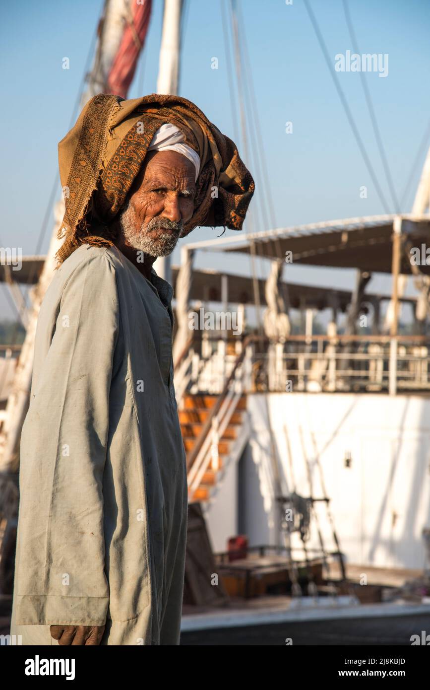 older Egyptian man on the bank of the Nile River with a Nile Dahabiya Boat in the background Stock Photo