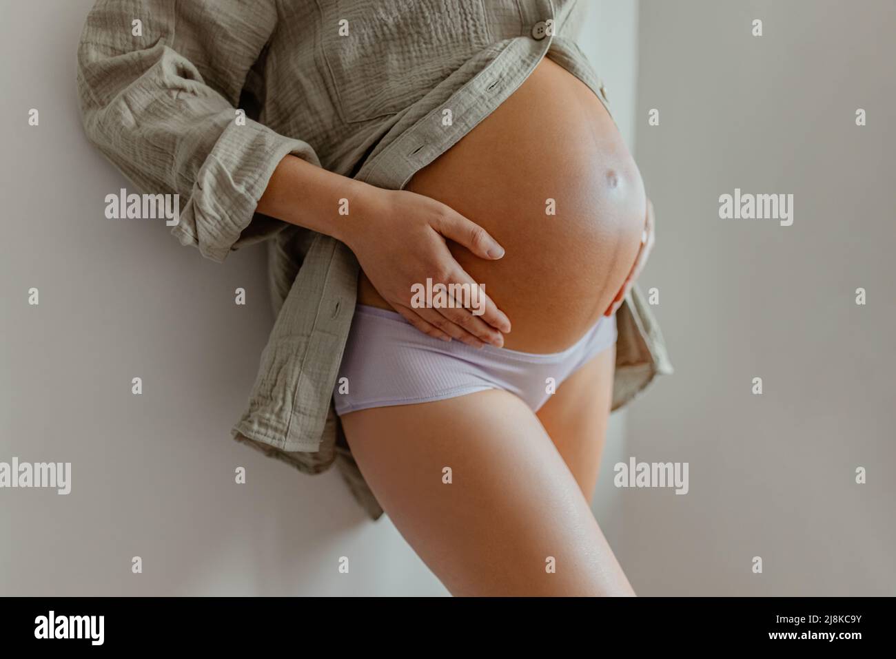 Pregnant woman wearing maternity underwear pajamas at home relaxing holding expecting tummy for skincare, health, lifestyle. Pregnancy belly closeup Stock Photo