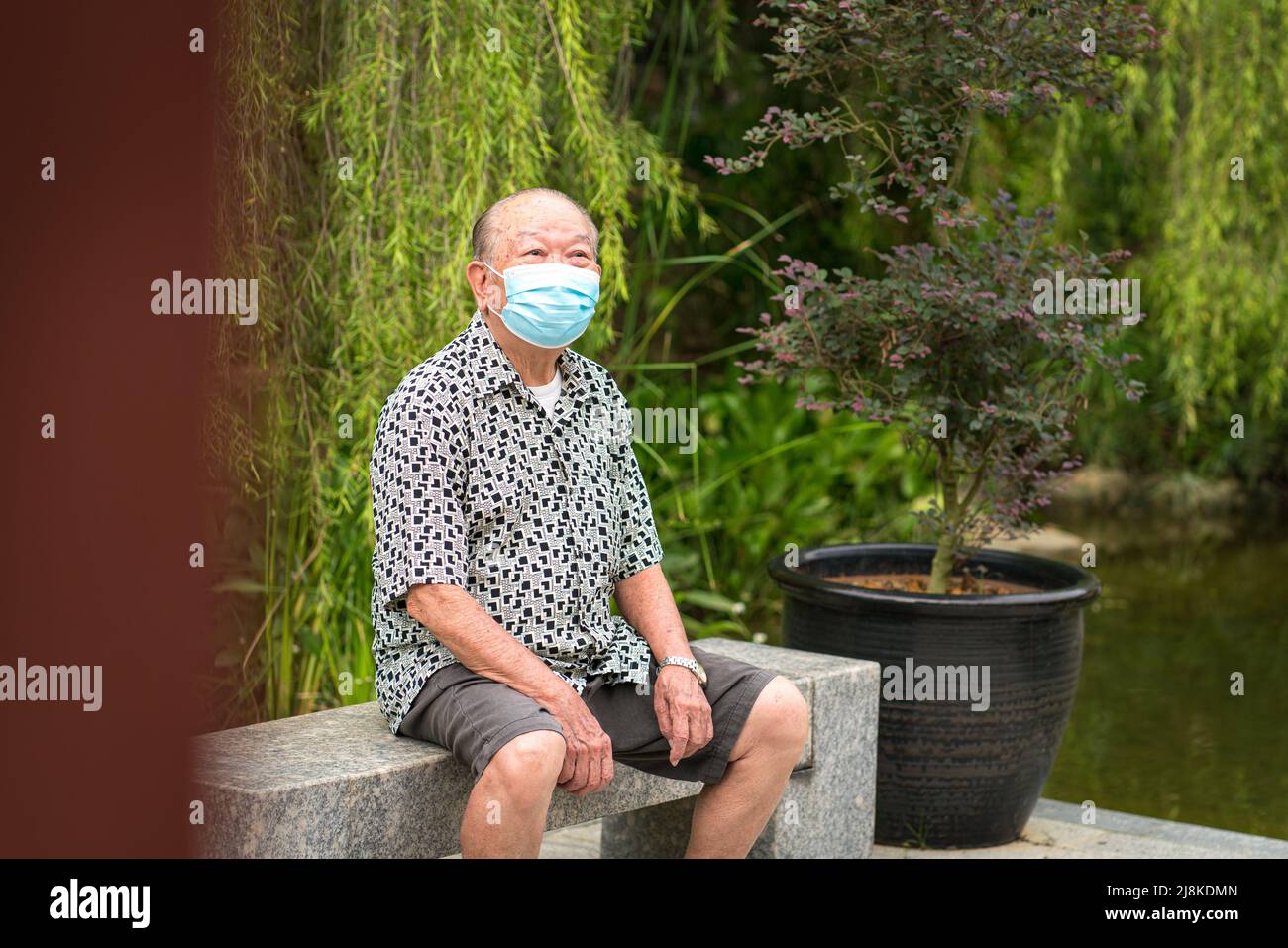 Senior Asian man with face mask on, sitting on a bench. Outdoor garden setting. Stock Photo