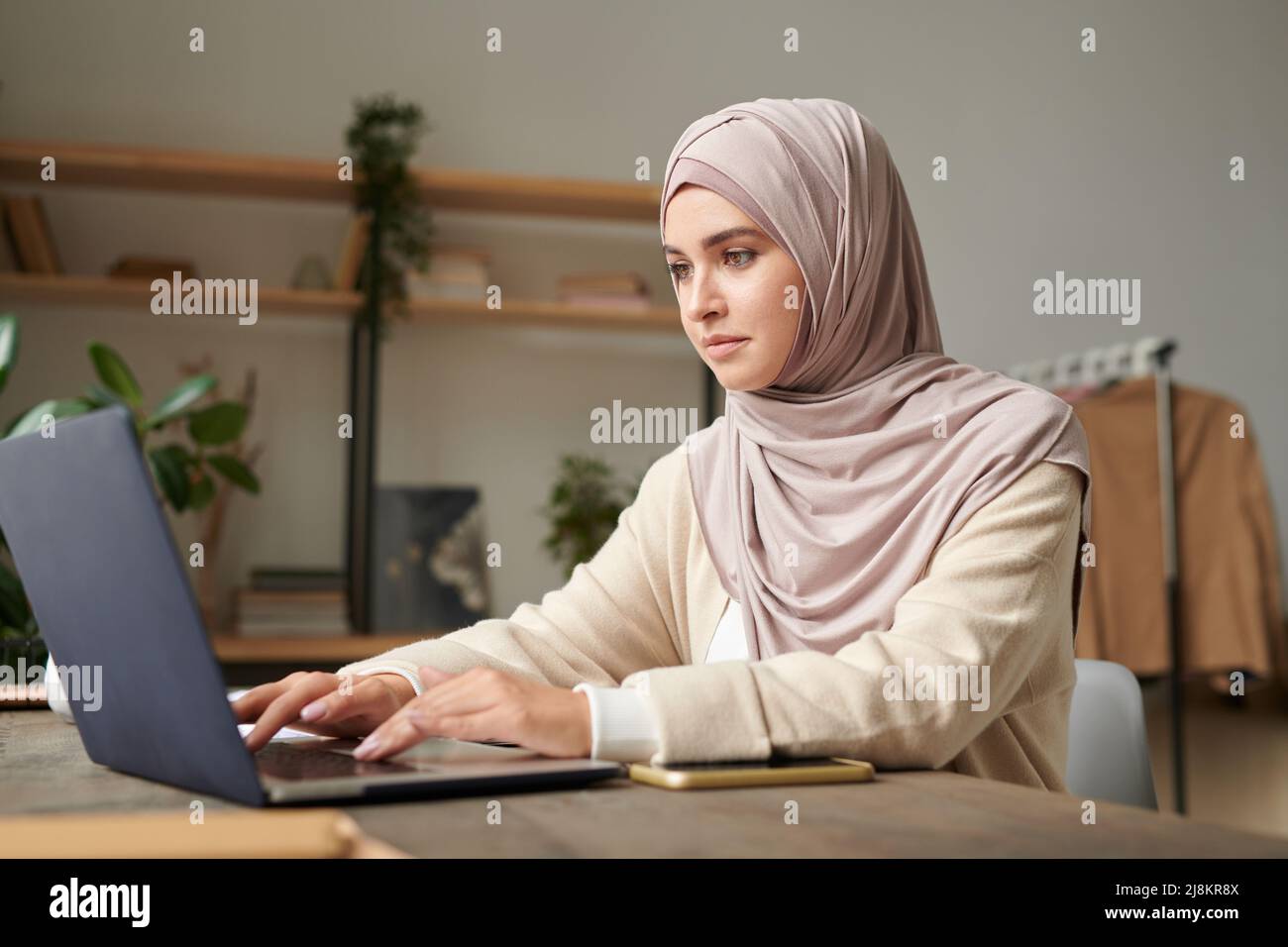 Horizontal portrait of attractive Middle Eastern woman wearing hijab spending time in modern office room working on laptop Stock Photo