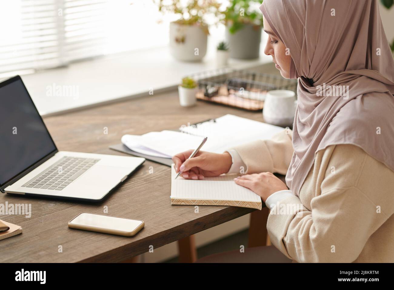 Successful Muslim woman wearing headscarf making notes in her copybook while working on business plan Stock Photo