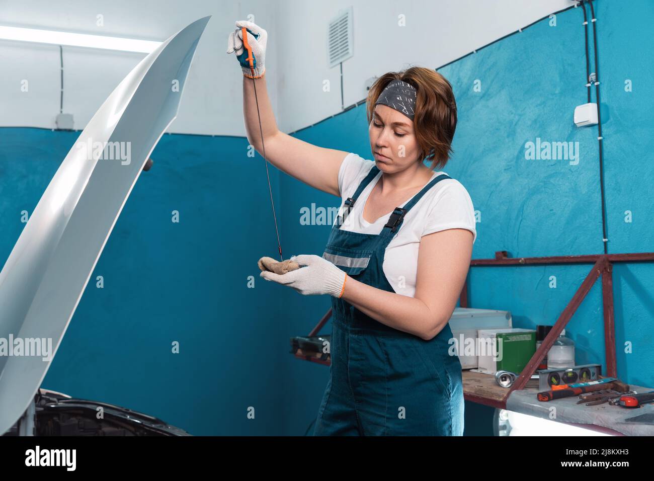 Young woman mechanic in blue overall and gloves repairs oil dipstick of ...