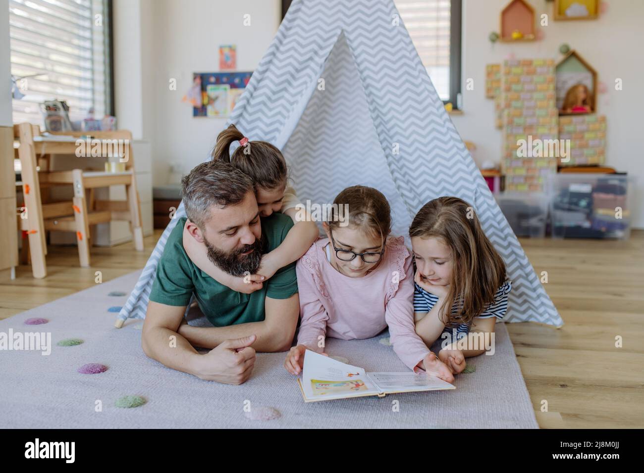 Cheerful father of three little daughters reading them book at home. Stock Photo