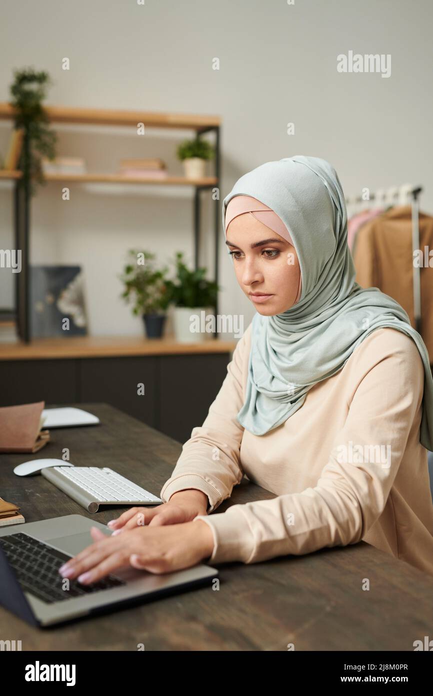 Vertical portrait of beautiful young woman wearing light blue hijab sitting at table working on laptop at home Stock Photo