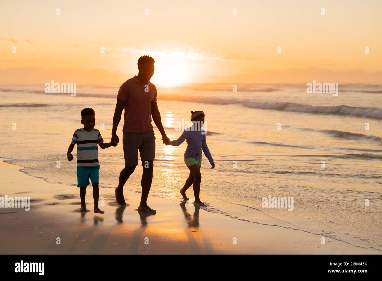 African american young father holding son and daughter's hands while walking on beach against sky Stock Photo