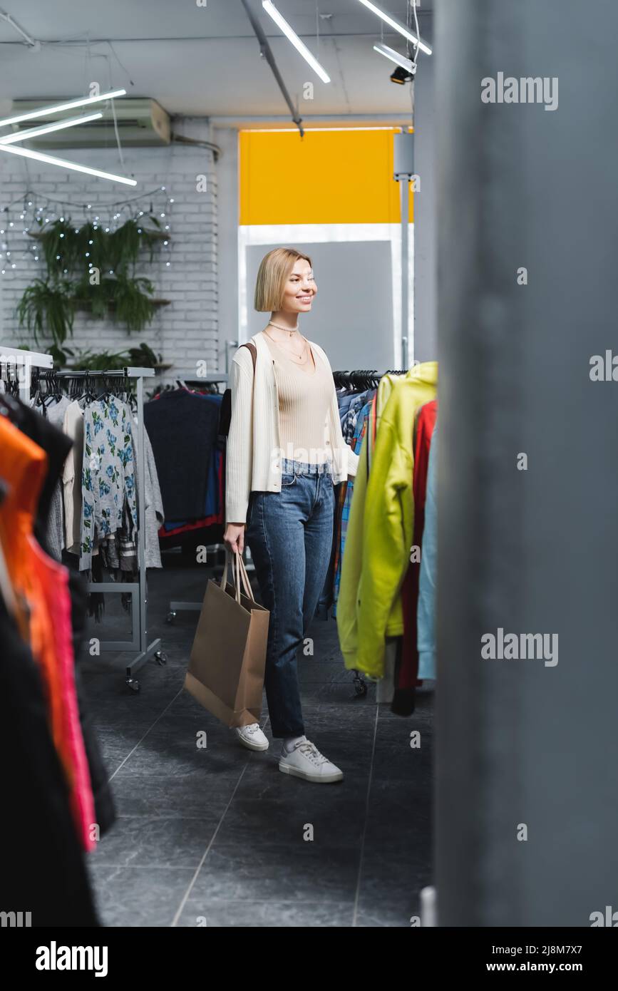 Positive blonde woman holding shopping bag near clothes on racks in second hand Stock Photo