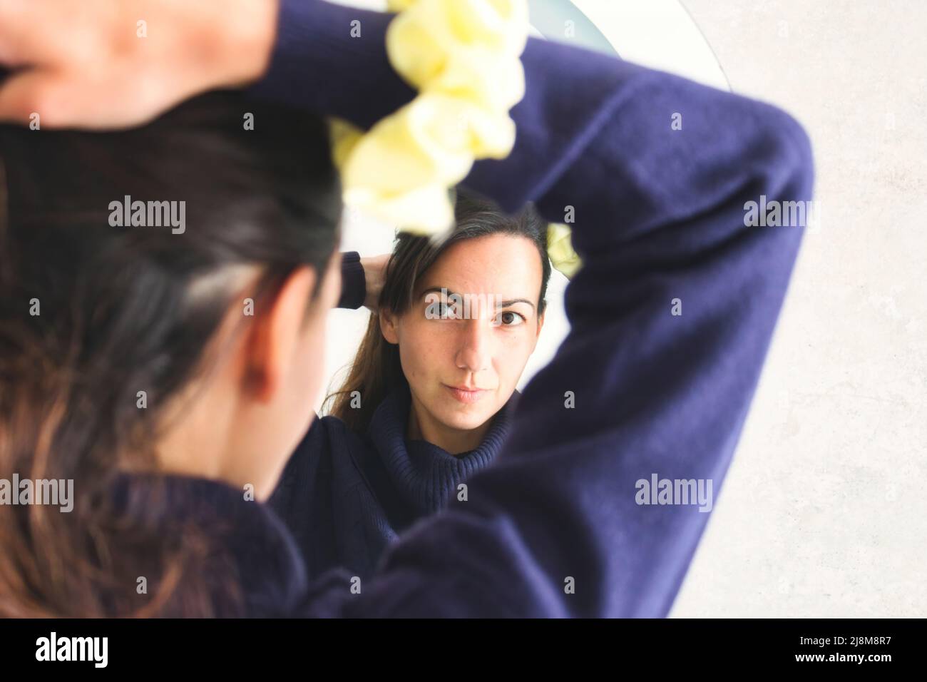 Young attractive woman looking at herself in the mirror and tying her hair into a ponytail with a scrunchie Stock Photo