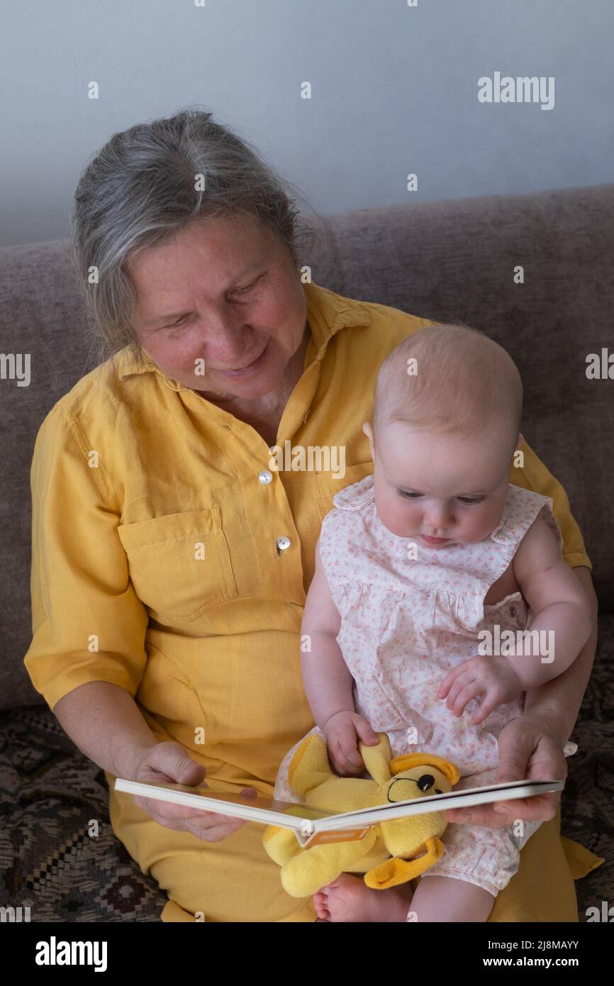 Loving grandmother reading a book to baby girl holding book sitting on sofa. Nanny granny telling story to a grandchild Stock Photo