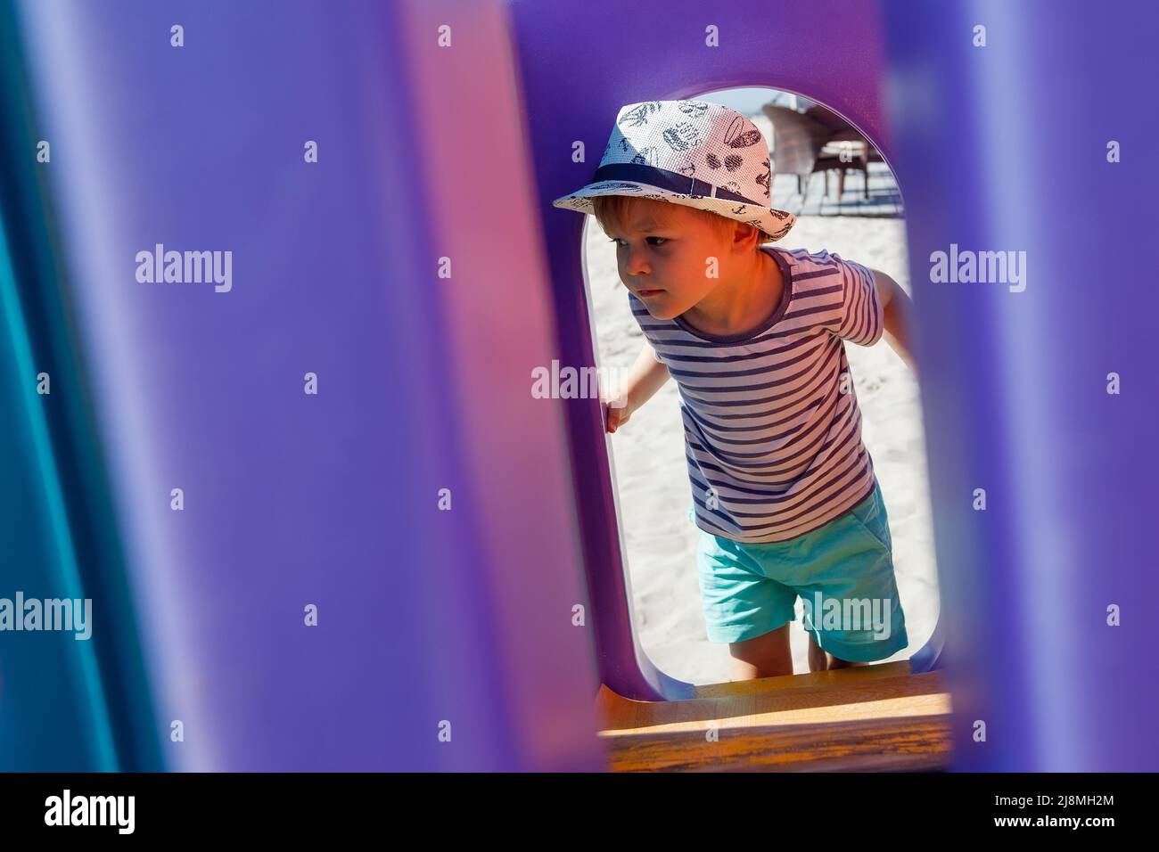A little boy in a hat looks at the playground through a purple plastic arch. The purple background has free space for the note Stock Photo