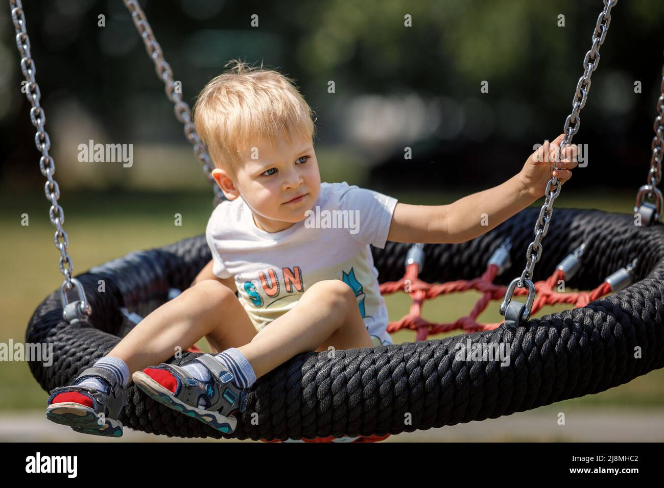A cheerful little boy swings in the park on a swinging rope swing. Round black and red seat for