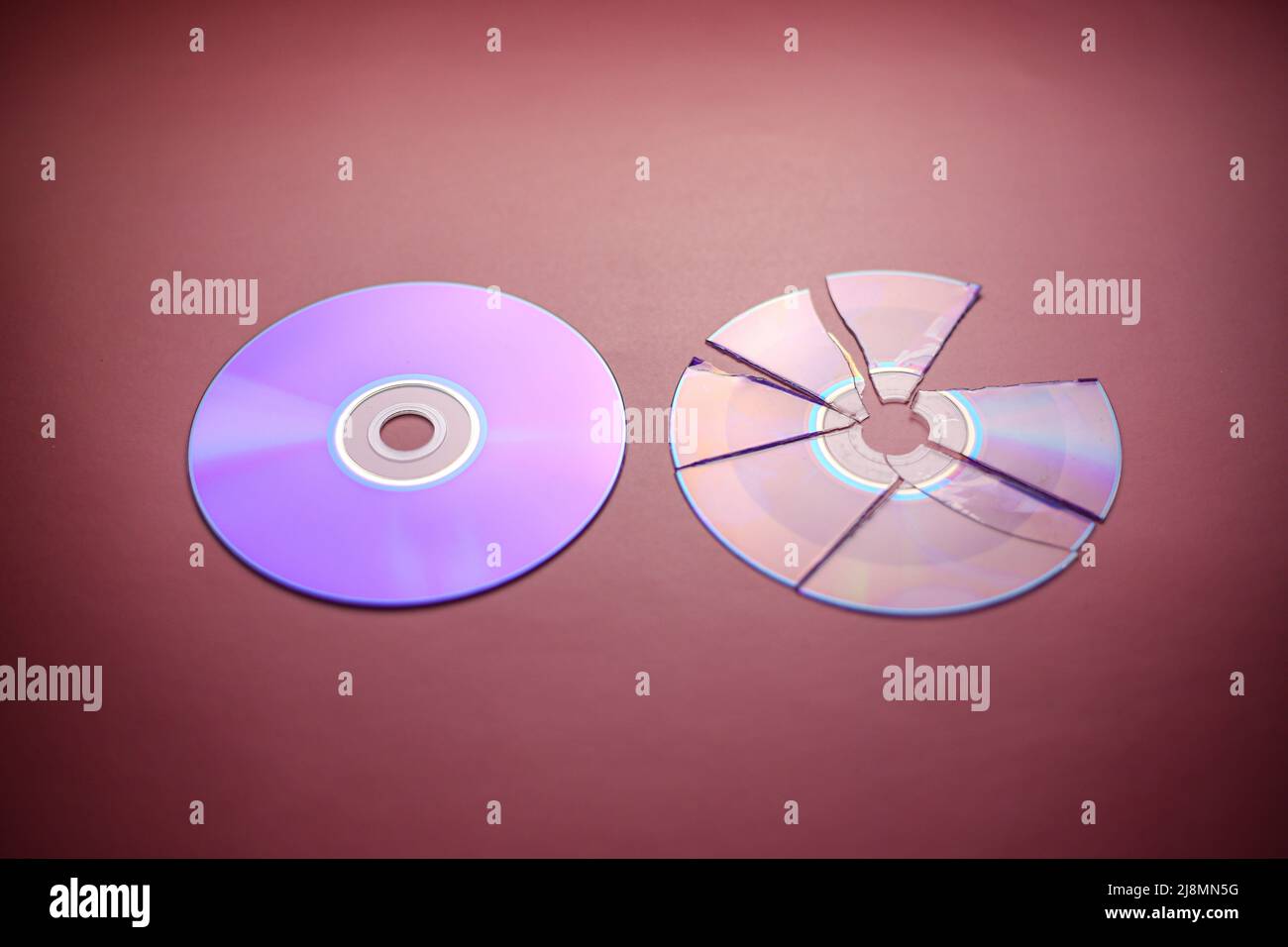 Broken compact disc divided into parts and a whole compact disc close-up on a red-burgundy background, complete loss of data Stock Photo