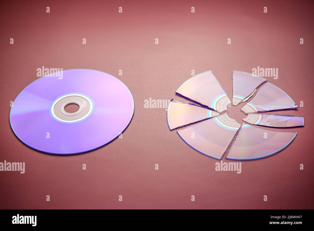 Broken compact disc divided into parts and a whole compact disc close-up on a red-burgundy background, complete loss of data Stock Photo