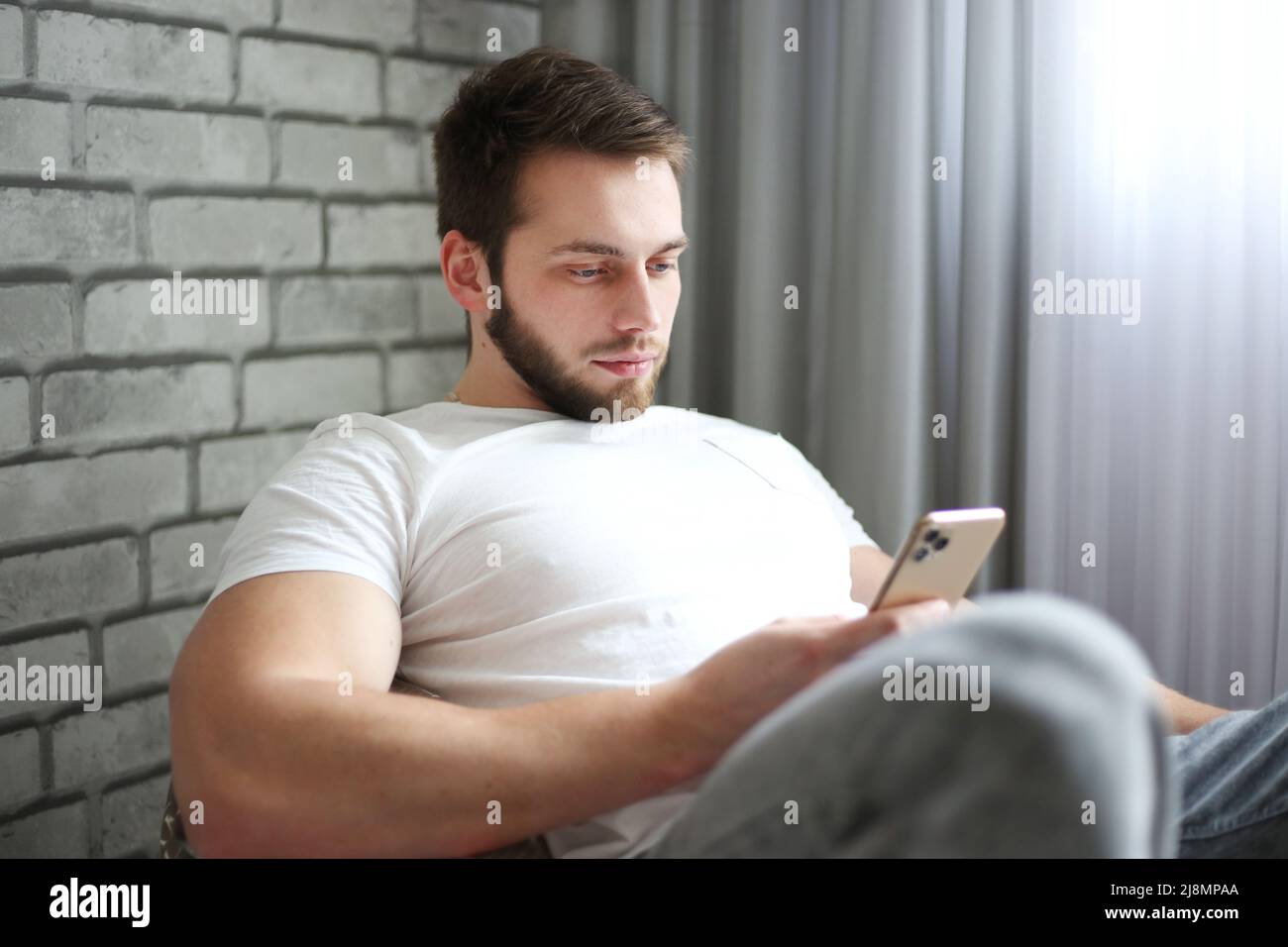 A young man of athletic build in a white T-shirt sits on a cushion chair and looks intently into smartphone Stock Photo