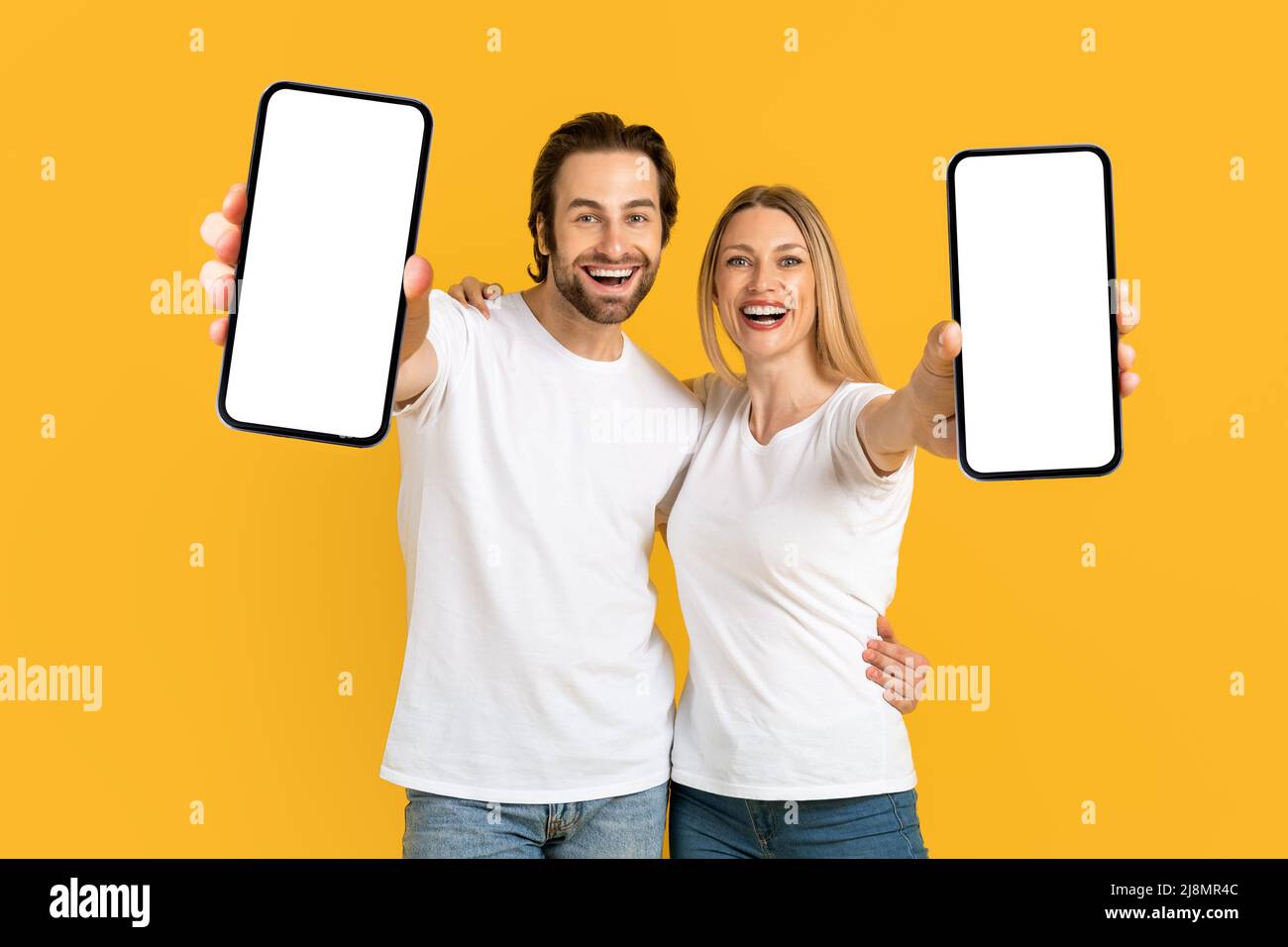 Cheerful young european wife and husband in white t-shirts show phones with blank screen Stock Photo