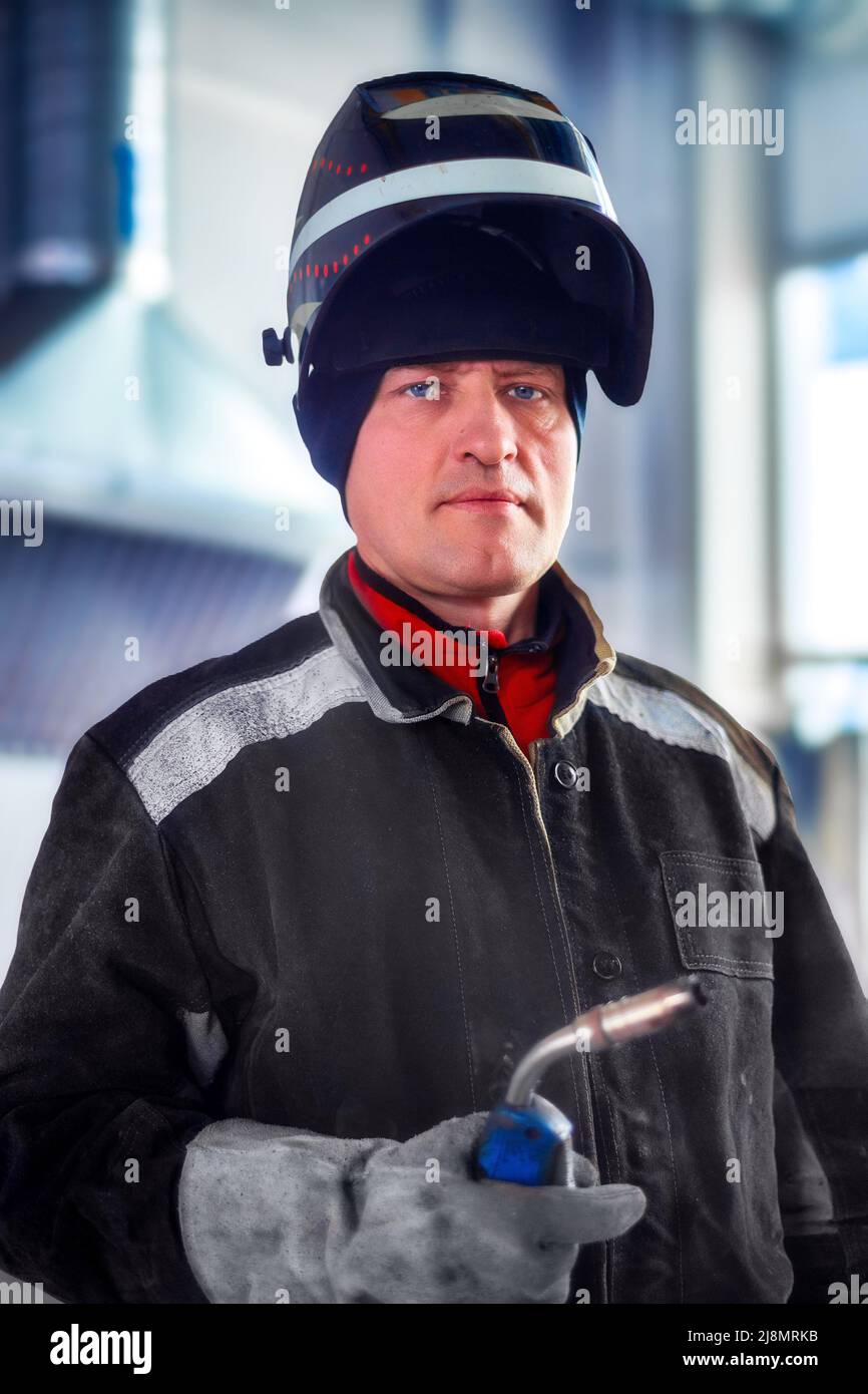 Male welder in work clothes with mask on his head looks directly into camera. Authentic portrait of worker in production hall. Real people. Stock Photo