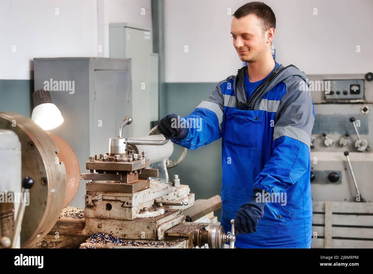 Young turner in overalls works on lathe in workshop. Authentic scene workflow. Caucasian worker recycles metal parts. Stock Photo